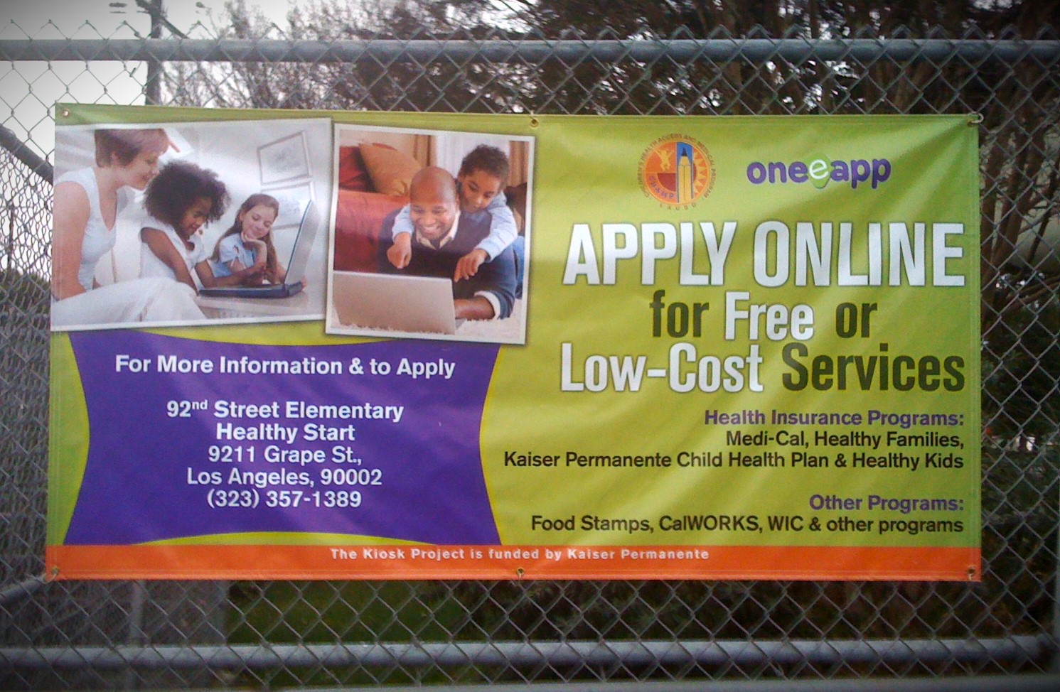 A large banner on a chain-link fence has images of children and adults looking at a computers and smiling. The headline reads, "One-e-App, Apply online for Free or Low-Cost Services." The subheading reads, " Health insurance programs; Medi-Cal, Healthy Families, Kaiser Permanente Child Health Plan & Health Kids. Below that it reads: Other programs: Food Stamps, CalWORKS, WIC, and other programs. On the left side of the banner, it says "For more information and apply..." and provides an address for an elementary school in Los Angeles." This is an old banner advertising the recently shut down app One-e-App